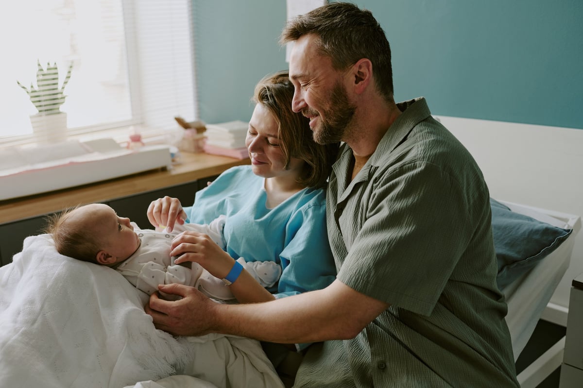 Caucasian parents holding newborn while sitting in hospital room embracing joyful moment and bonding with infant on bed with cozy room